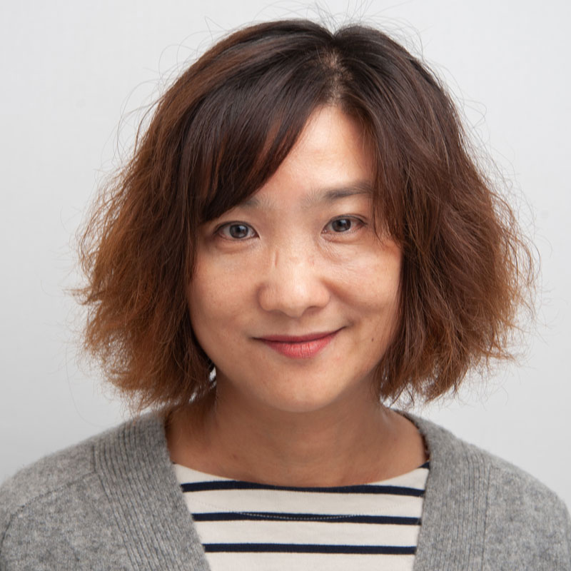 Image of a woman light brown skin with short brown/auburn hair and brown eyes. She is wearing a black and white stripped shirt with a grey jumper over the top.
