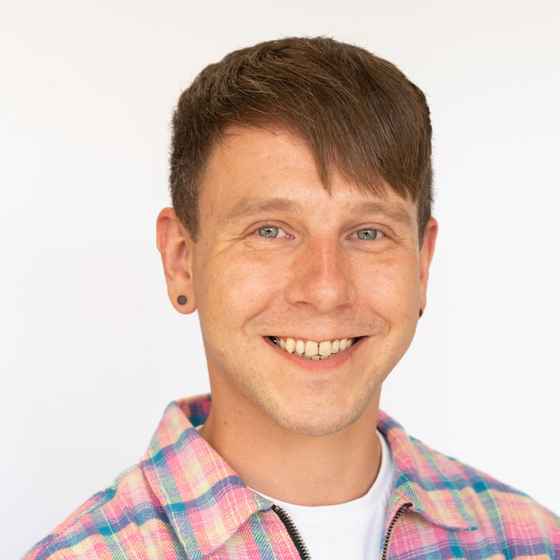 Image of a white man with light brown hair and light green eyes. He is wearing brown stud earrings and a pastel stripped jacket over a white t-shirt.
