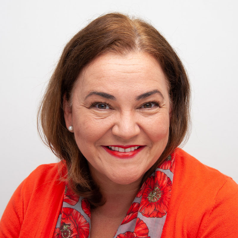 Image of a woman with light brown skin, auburn wavy should length hair and brown eyes. She is wearing pearl stud earrings, an orange cardigan and a scarf with orange poppies on it.