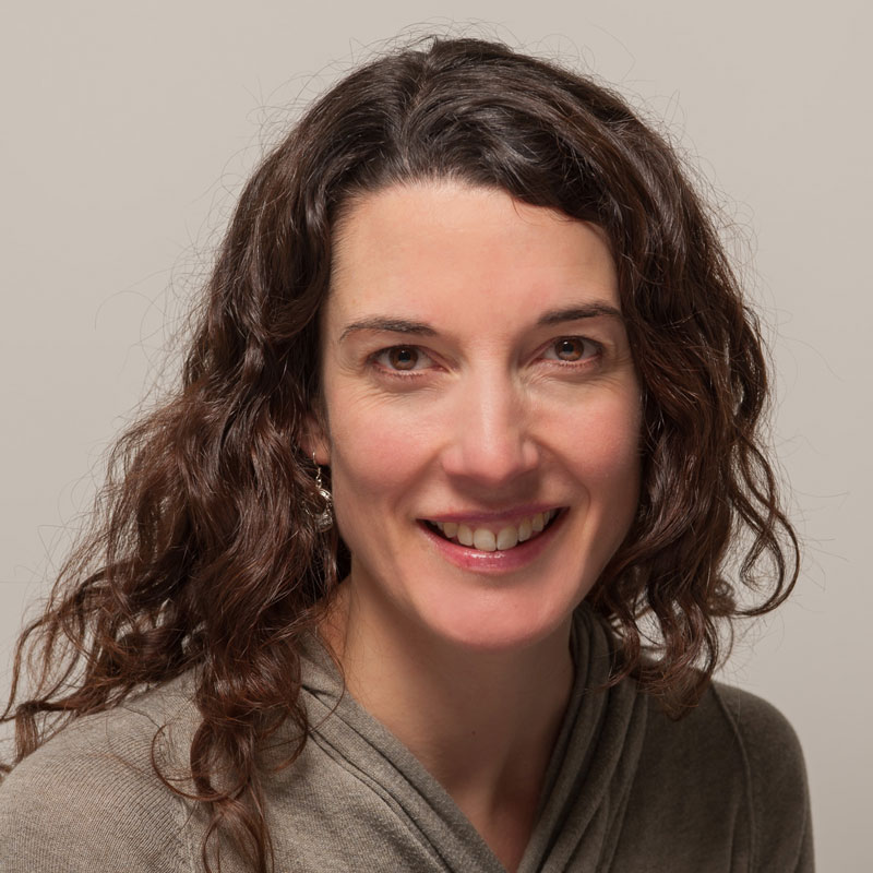 Image of a white woman with long dark brown, curly hair, brown eyes and and beige top.