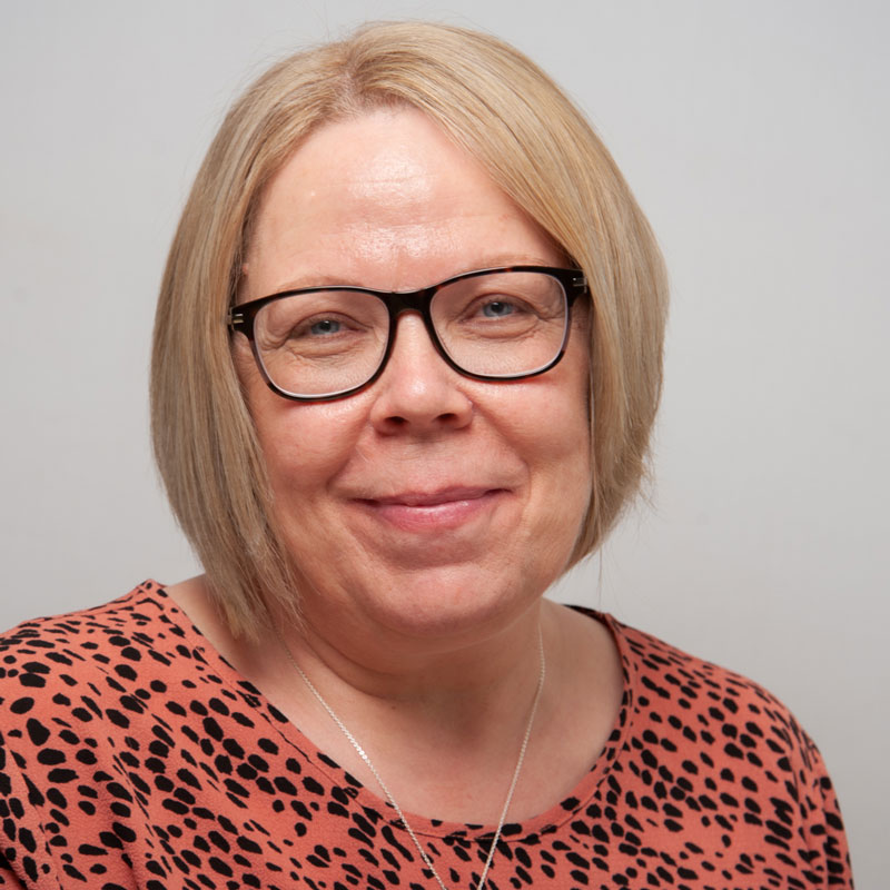 Image of a white woman with light blonde straight hair cut in a short bob. She has light blue eyes and is wearing black rectangular glasses. She is wearing a pink top with black spots and a necklace with a silver chain.