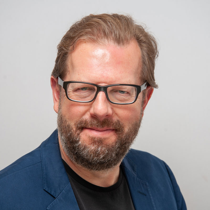Image of a white man with dark blonde/light brown hair and a dark brown/grey short heard. He has blue eyes and is wearing black framed glasses and a navy blue blazer and a black t-shirt.