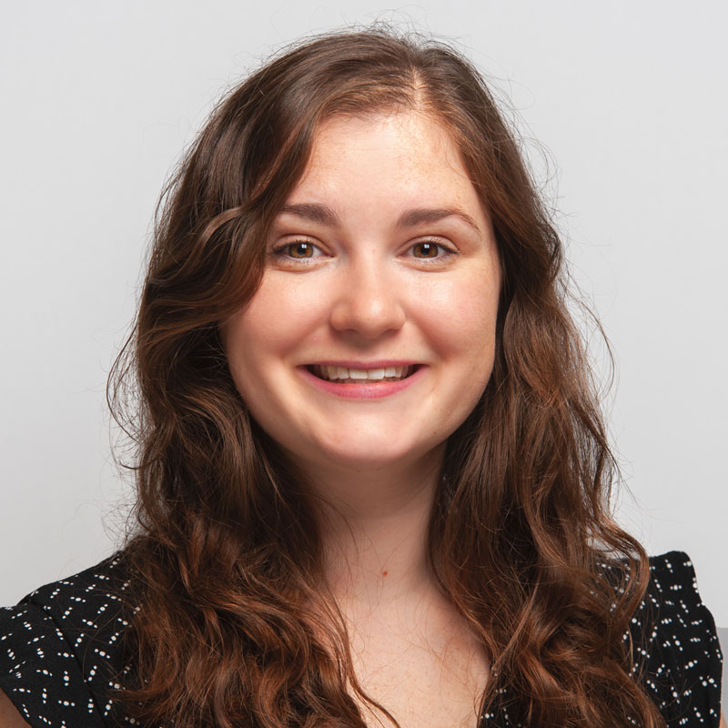 A white woman with long brown/auburn wavy hair and hazel eyes. She is wearing a black top with white dots on it.