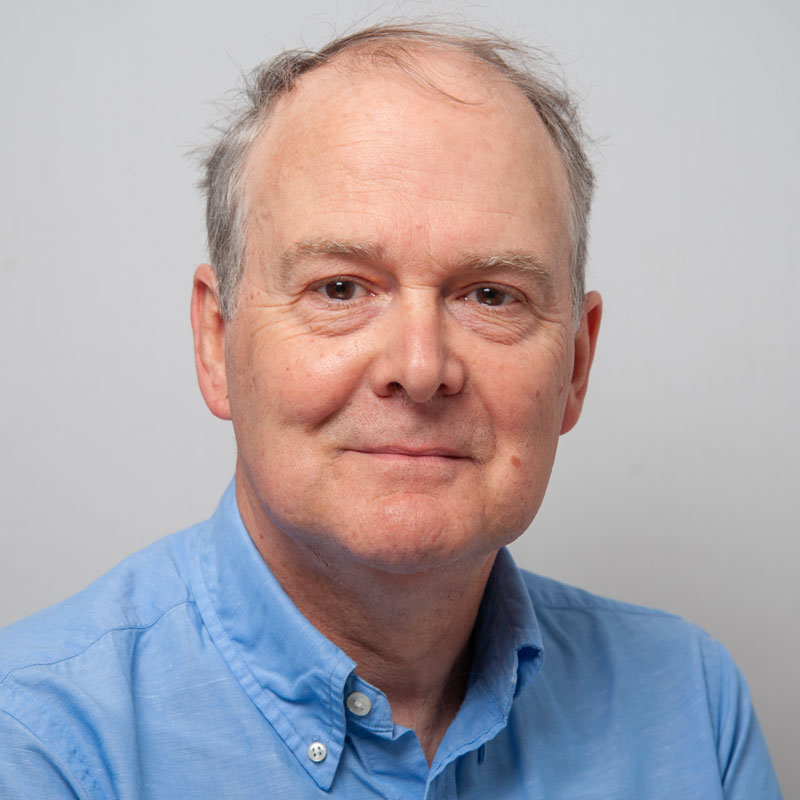 Image of a white man with grey hair around the sides of his head, light brown eyes and a blue collared shirt.