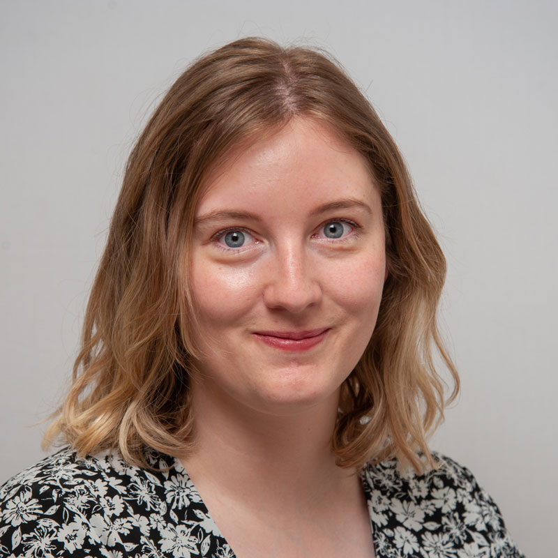 Image of a white woman with medium blonde wavy shoulder length hair. She has light blue eyes and is wearing a black shirt with a white floral design.