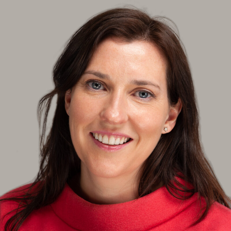 Image of a white woman with dark brown long hair, blue eyes and star stud earrings. She is wearing a coral cowl neck jumper.