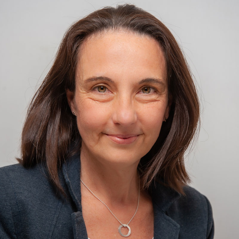 Image of a white woman with medium brown straight hair and light brown eyes. She is wearing a slate blue blazer and a silver necklace with a round ring on it.
