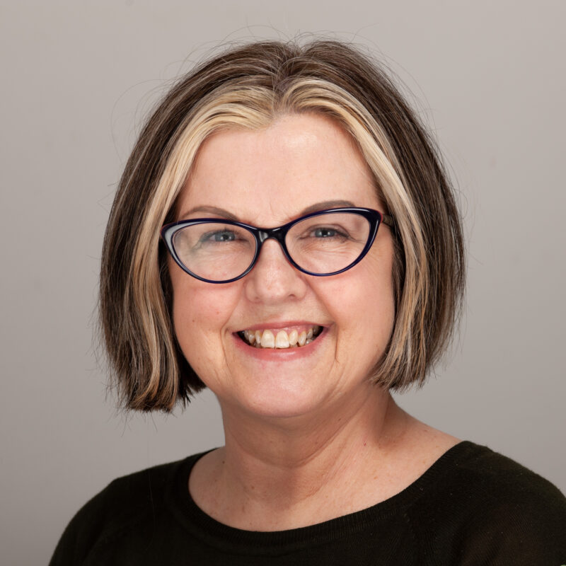 Image of a white woman with brown hair with chunky blonde highlights cut in a bob. She is wearing black cat eye glasses and a black shirt.