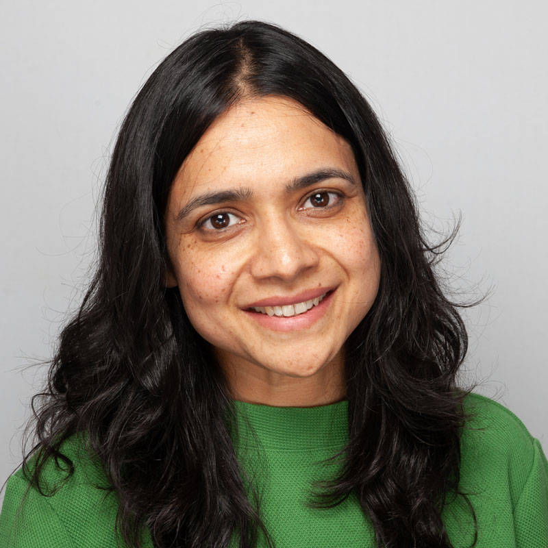 Image of a woman with light brown skin with long black wavy hair and brown eyes. She is wearing a grass green jumper.