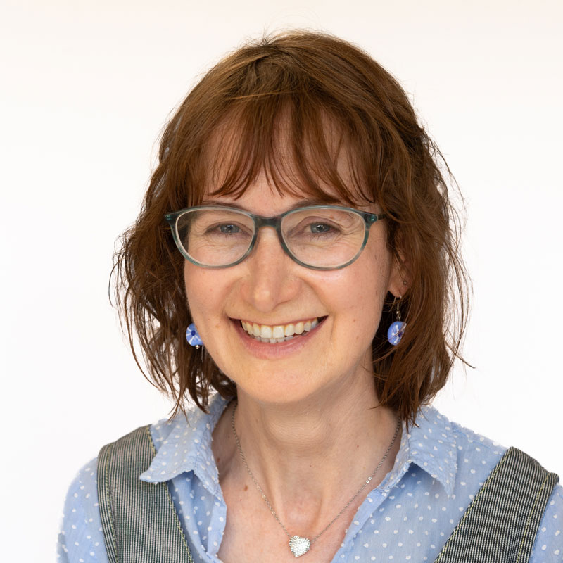 Image of a white woman with shoulder length auburn wavy hair with a fringe. She has hazel eyes and is wearing sage green glasses. She is wearing light blue drop earrings, a silver heart necklace and a light blue polka dot collared shirt.