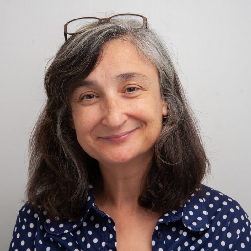 Image of a white woman with olive skin, dark brown and grey hair and brown eyes wearing a blue and white polka dot shirt and brown glasses on top of her head.