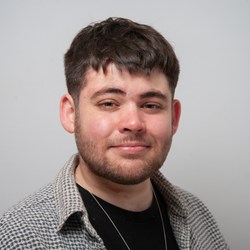 Image of a white man with dark brown hair, blue eyes and a brown beard. He is wearing a black t-shirt with a silver chain necklace and a checked black and white collared shirt on top.