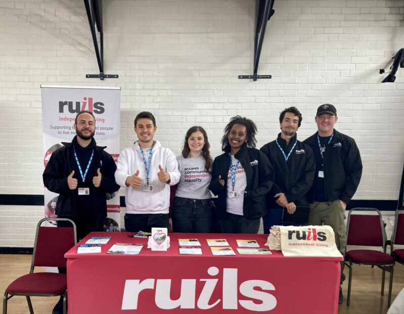 Image of 4 men and 2 woman standing behind a table with a Ruils table cloth on it. Two of the men have their thumbs up.