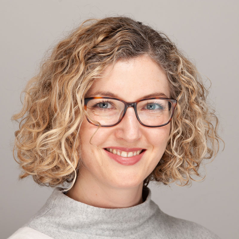 Image of a a white woman with light green eyes and short blonde curly hair. She is wearing brown rectangular glasses and a grey high necked jumper.