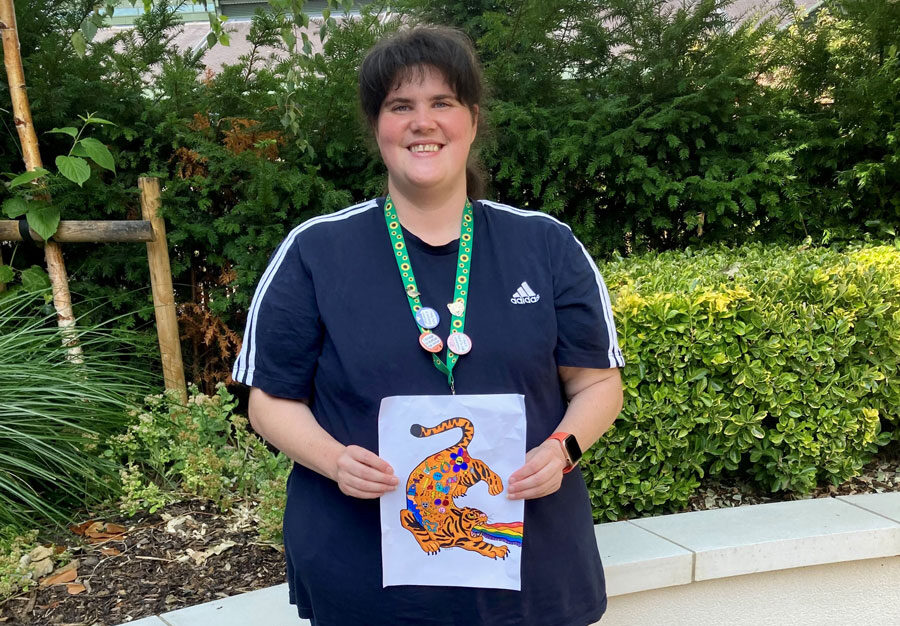 Image of Social Prescribing client Emma holding a piece of original artwork. She is standing in front of a stone wall with hedges behind her.