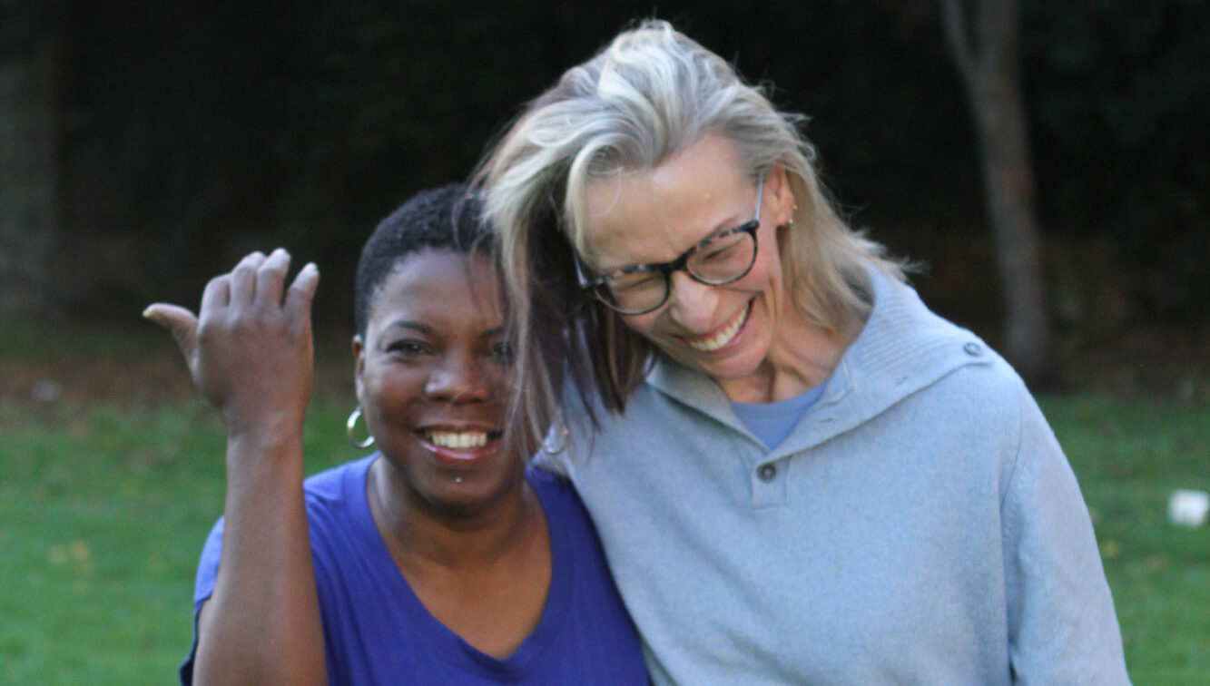 Image of a client and her personal care assistant the client is wearing a light blue jumper and the personal care assistant is wearing a dark blue T shirt. Both are laughing and facing the camera.