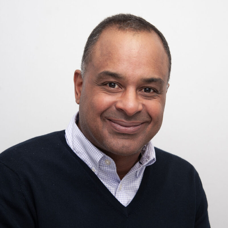 Image of a black man with short brown and white hair and brown eyes. He is wearing a blue and white checked shirt under a navy blue jumper.