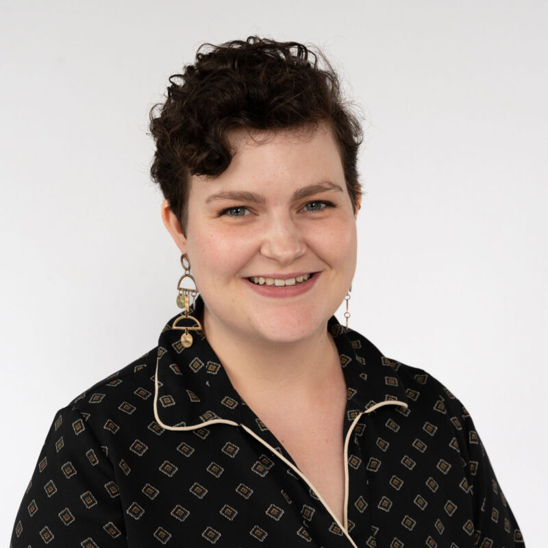 Image of a white woman with short dark brown curly hair and blue eyes wearing gold dangly earrings and a black shirt with a gold geometric pattern.