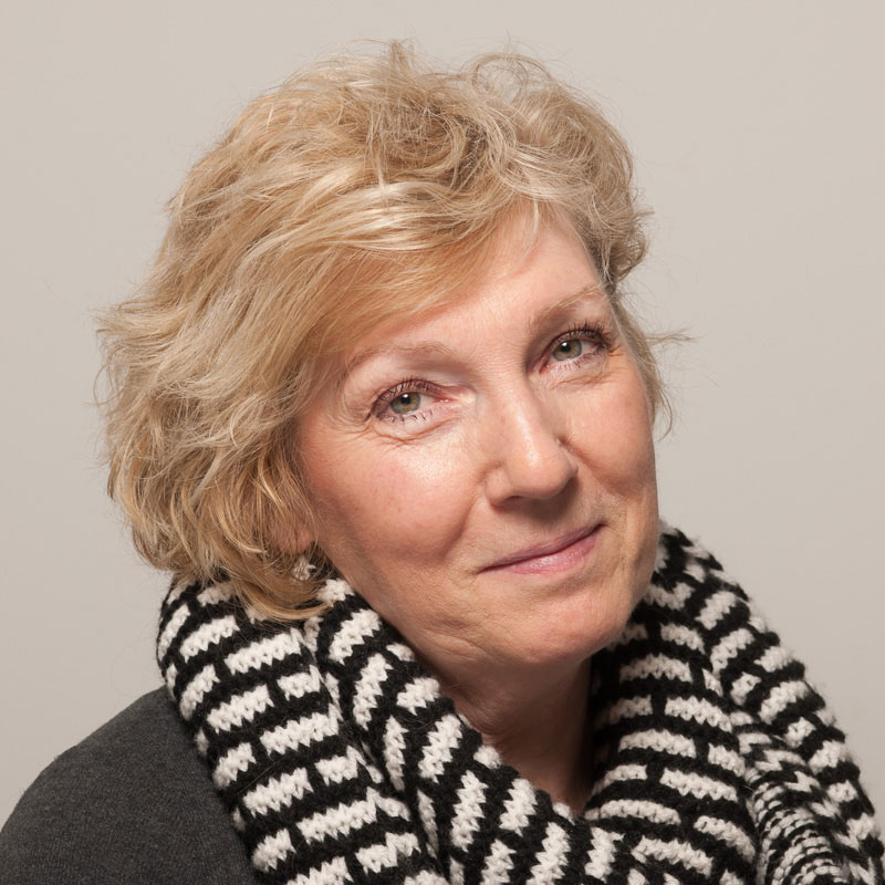 Image of a white woman with short light blonde hair, hazel/green eyes wearing a black and white chunky scarf over a grey top.