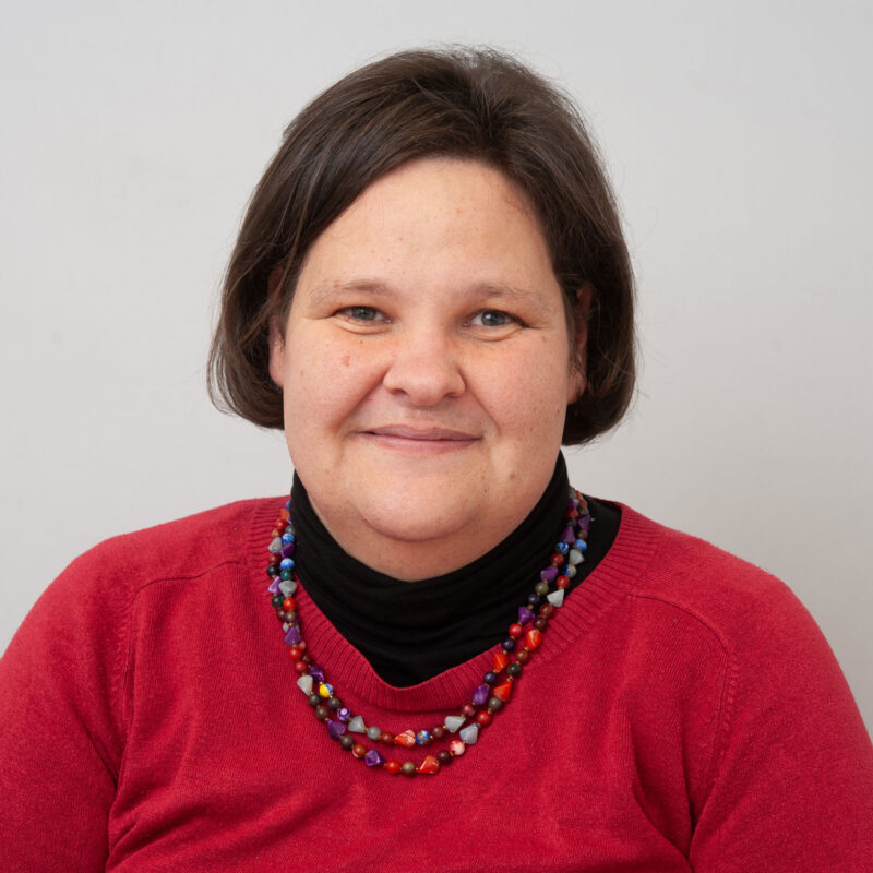 Image of a white woman with dark brown hair cut in a bob with light brown eyes. She is wearing a red jumper over the top of a black turtleneck and a purple and red beaded necklace.