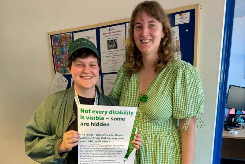 Two women, Dom and Jenny, standing holding a Hidden Disabilities Sunflower poster that reads "Not every disability is visible, some are hidden". Both are wearing green clothes.