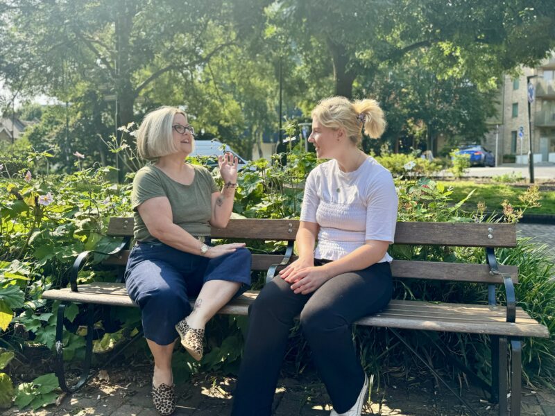 Two women sitting on a wooden bench in a town square speaking with one another. Behind them are trees and shrubs.