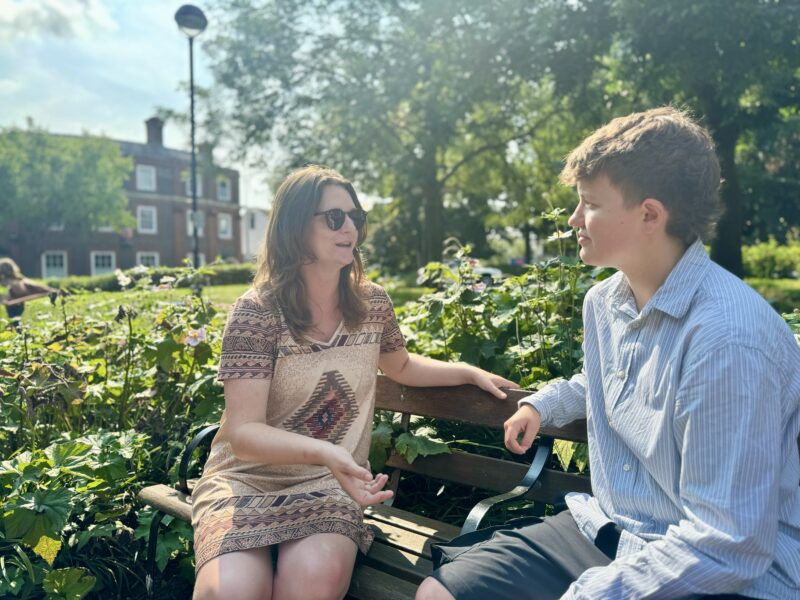 Two women sitting on a park bench talking to each other. One is wearing sunglasses and gesturing to the other.