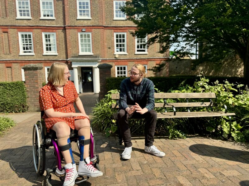 Image of a female wheelchair user in a red dress chatting with a man sitting on a park bench in a town square.