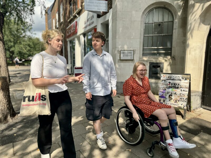 Three people going down the street talking with each other. One is wheeling herself in a wheelchair and the other two are walking. One has a Ruils tote bag on her arm.