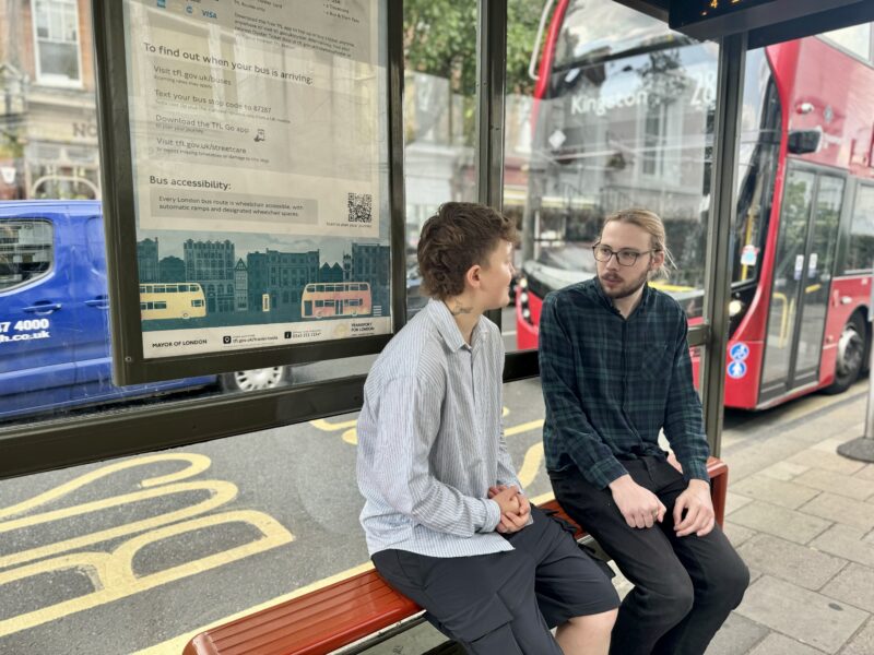 Two people sitting at a bus stop on Teddington high street. Behind them a red bus is going by.