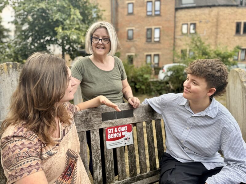 Image of two women sitting on a "sit and chat" bench and another woman standing behind the bench.