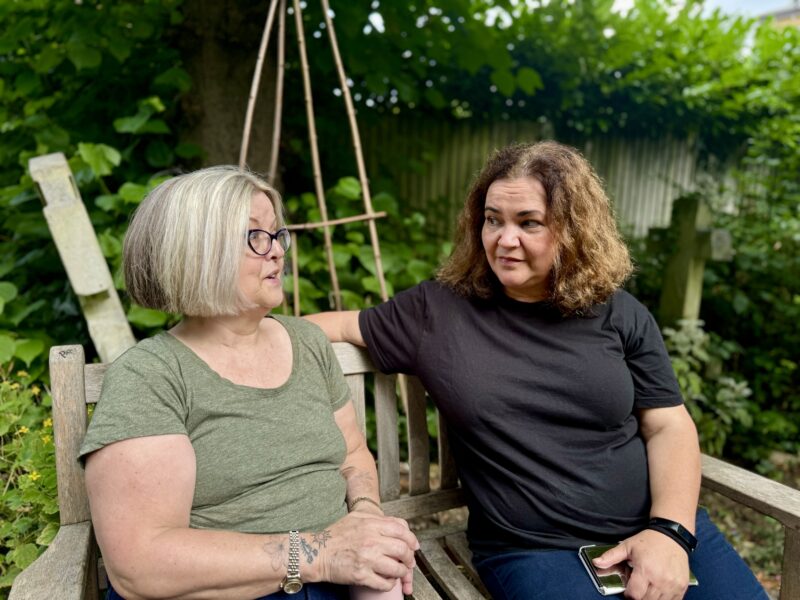 Two women sitting on a park bench in a community garden talking with each other.