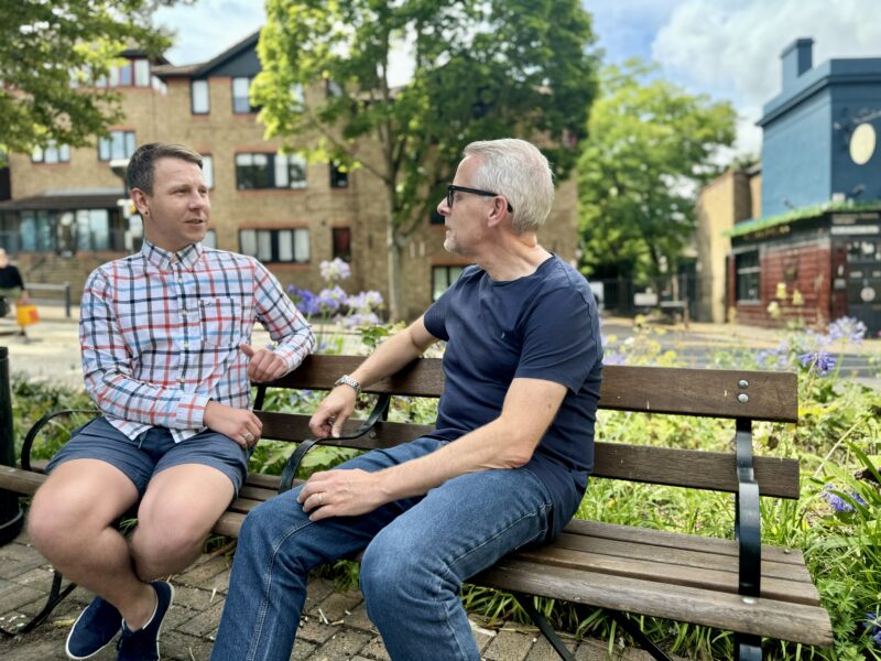 Two men sitting on a wooden bench in a town square speaking with each other.