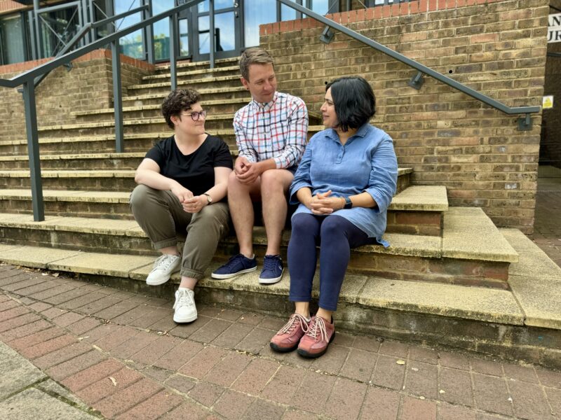 Three members of the Ruils team sitting on the steps at the entrance to an office building. Dom and Callum are listening to Surabhi speak.