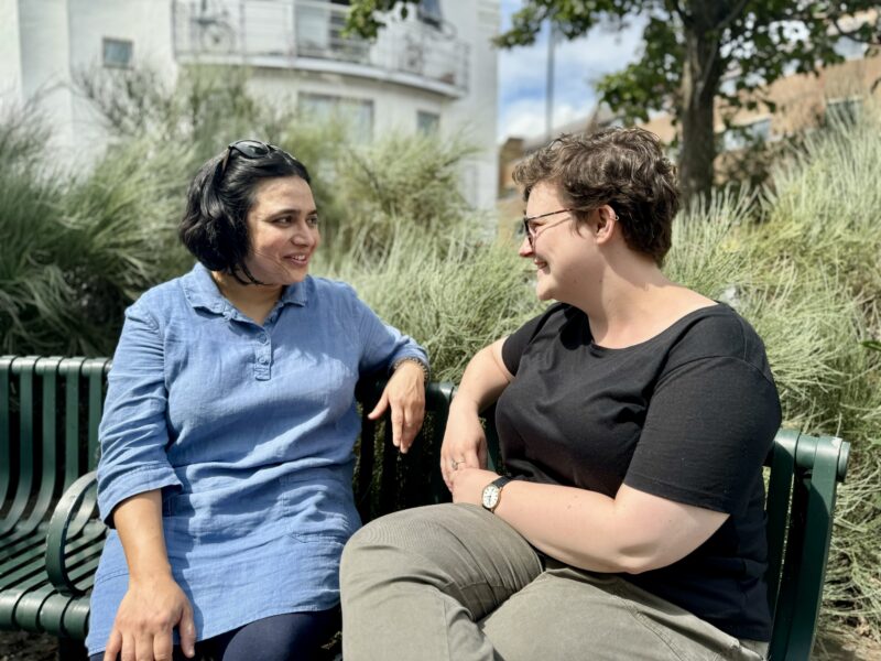 Two women sitting on a green bench in a park.