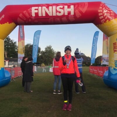 Image of CEO Cathy Wearing a bright orange jumper standing at the finish line of the 100 kilometre Ultra challenge walk.