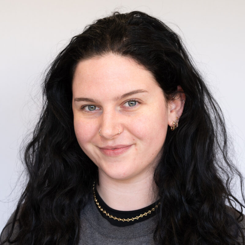 Image of a white woman with a fair complexion with black hair and blue eyes wearing a dark grey top. She has two nose piercings, ear piercings and is wearing a gold chain necklace.