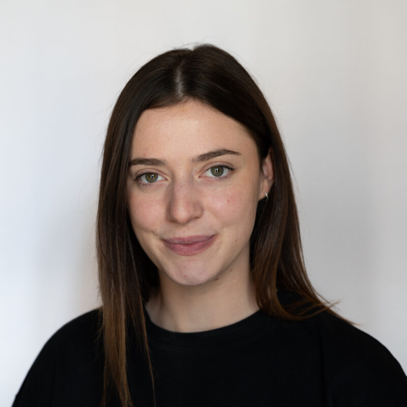 Image of a white woman with grey/green eyes and long dark brown hair wearing a black top.