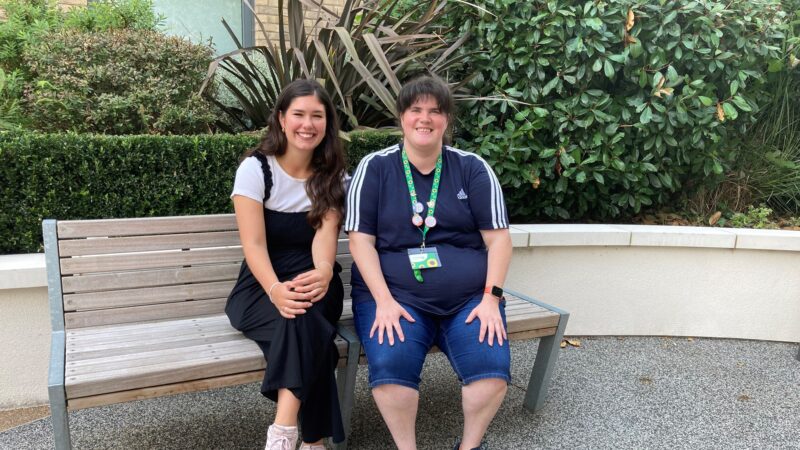 Image of a social prescriber Esther sitting with her client Emma who is faring a blue Adidas top and a sunflower land yard. They are seated on a bench and behind them is a hedge.