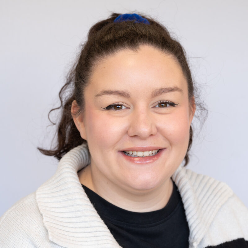 Image of a white woman with dark brown hair tied up in a ponytail and brown eyes. She is wearing a black shirt and a cream jumper.