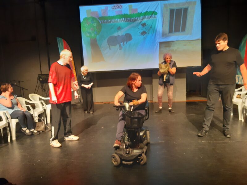 Image of the Ruils Theatre group performing at the Joy Festival in Hammersmith. They are on a stage with a black floor and have photos of drawings projected on a screen behind them.