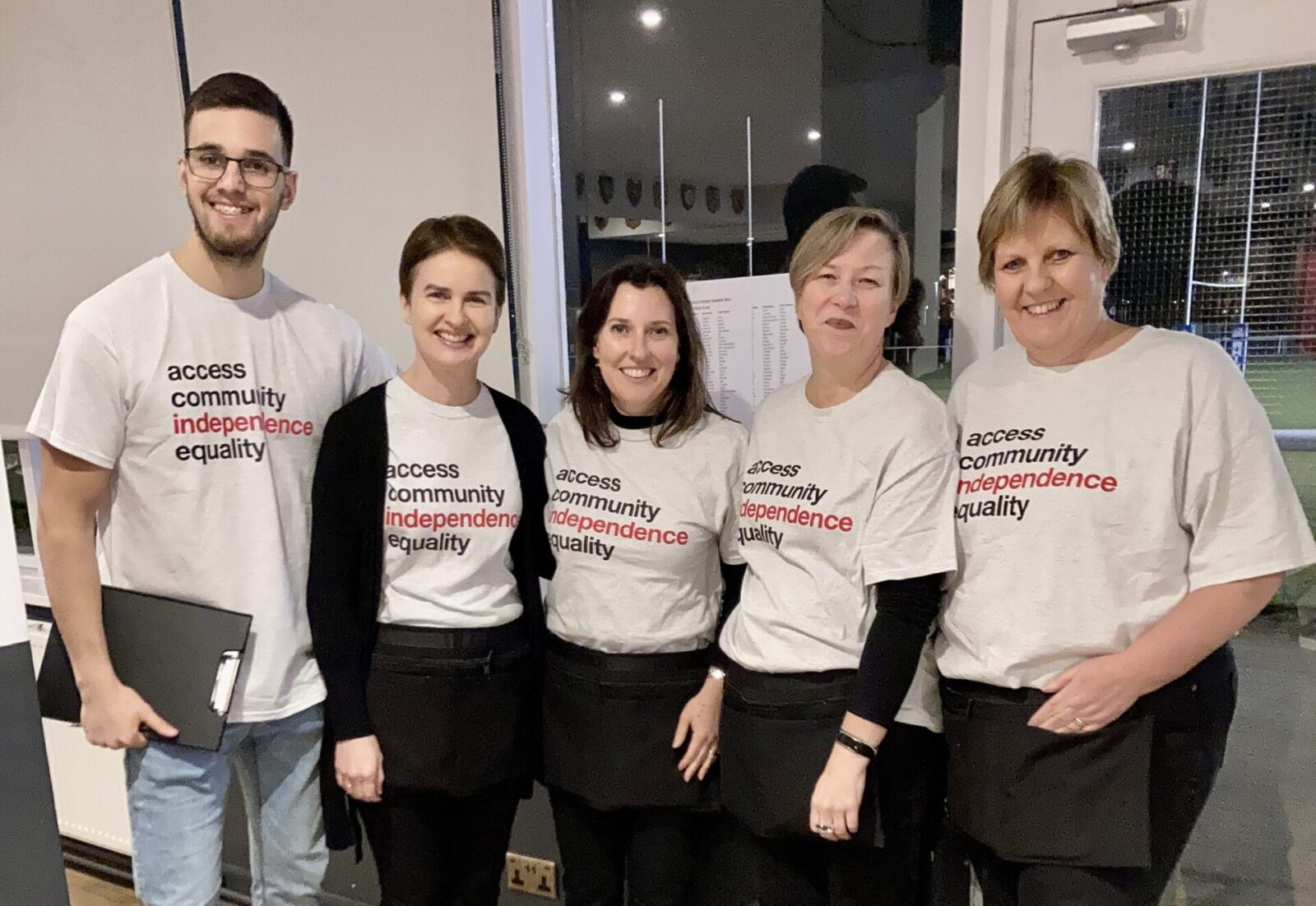 Image of five Ruils volunteers at the recent rugby dinner. Are wearing grey Ruils t shirts and standing in front of a white door.