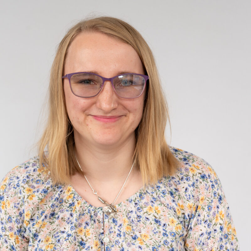 Image of a white woman with green eyes and blond shoulder length hair. She is wearing purple glasses, a silver necklace and a floral top.