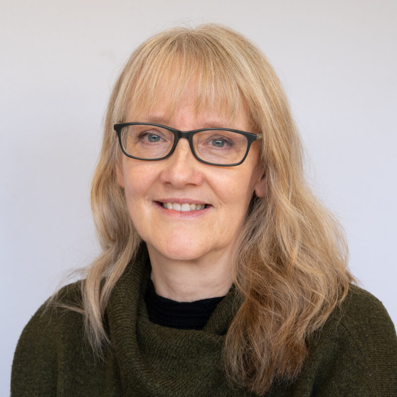 Image of a white woman with light blonde medium length hair with a fringe, wearing black rectangular framed glasses and a dark green jumper.