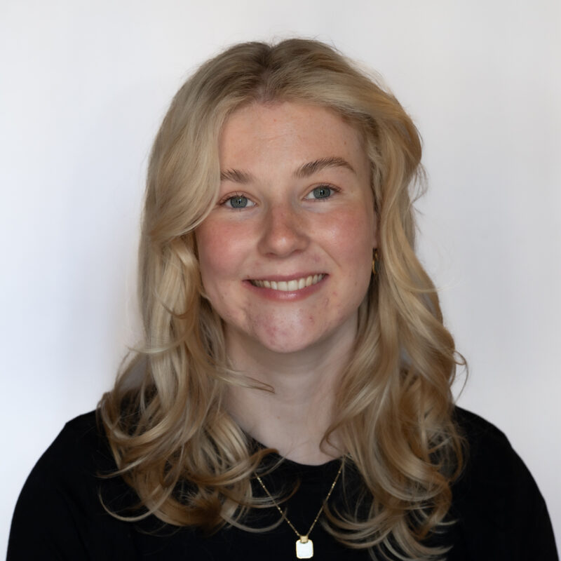 Image of a white woman with light blonde long hair, blue eyes and fair skin. She is wearing a black shirt and a silver and gold pendant necklace.