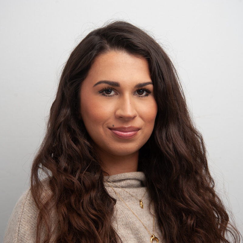 A woman with light brown complexion, long dark brown wavy hair, brown eyes wearing two gold necklaces and a tan jumper.