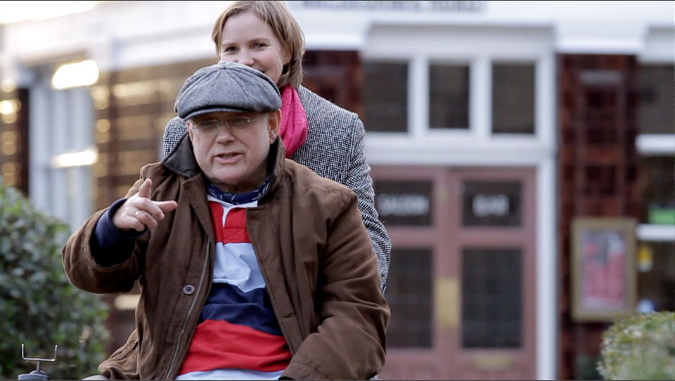 Image of a man wearing a grey flat cap in a wheelchair being pushed by a woman in a dark grey coat.