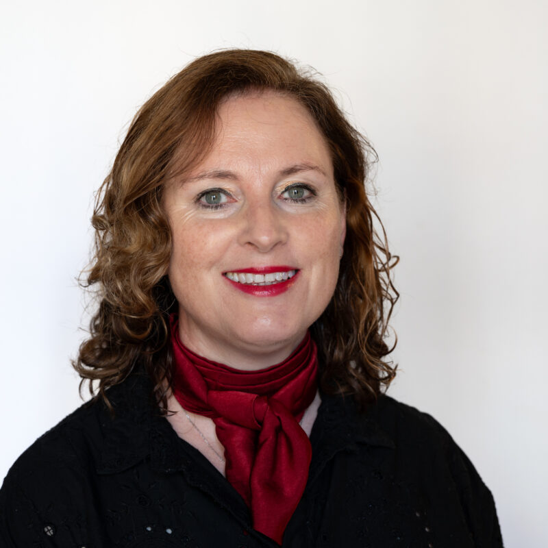 Image of a white woman with auburn hair and green eyes wearing a scarlet red neck scarf and a black top. She is wearing red lipstick.