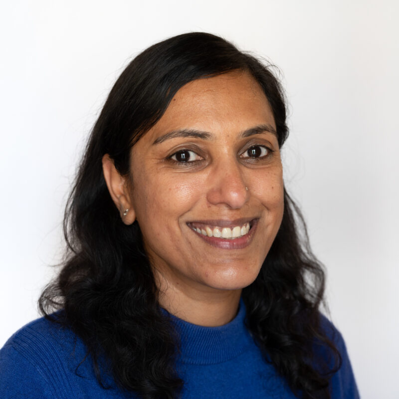 Image of a brown woman with black hair and brown eyes wearing a bright blue jumper. She has a nose piercing and is wearing silver stud earrings.