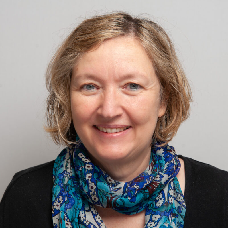 Image of a white woman with blue eyes and short medium blonde hair wearing a blue and white flower scarf and a black top.